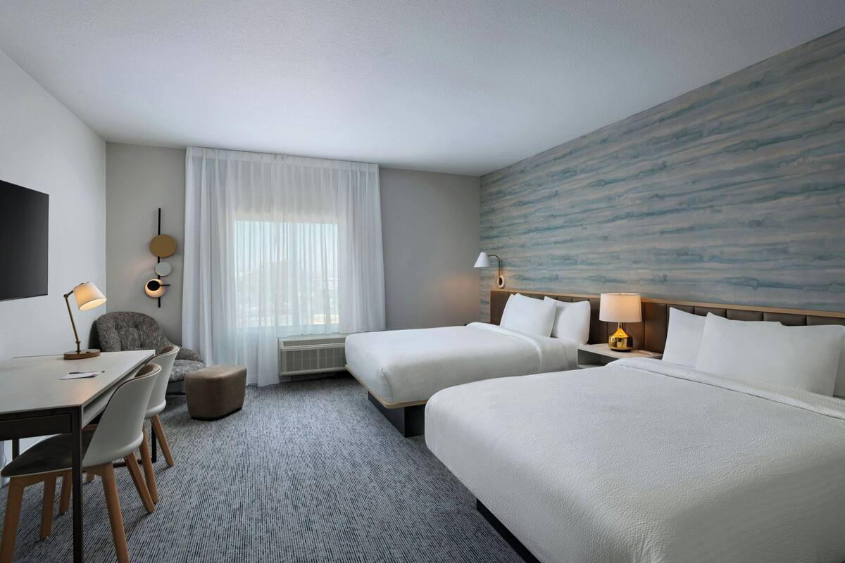 Une chambre d’hôtel avec deux lits, un bureau, des chaises, une lampe et une grande fenêtre avec des rideaux.