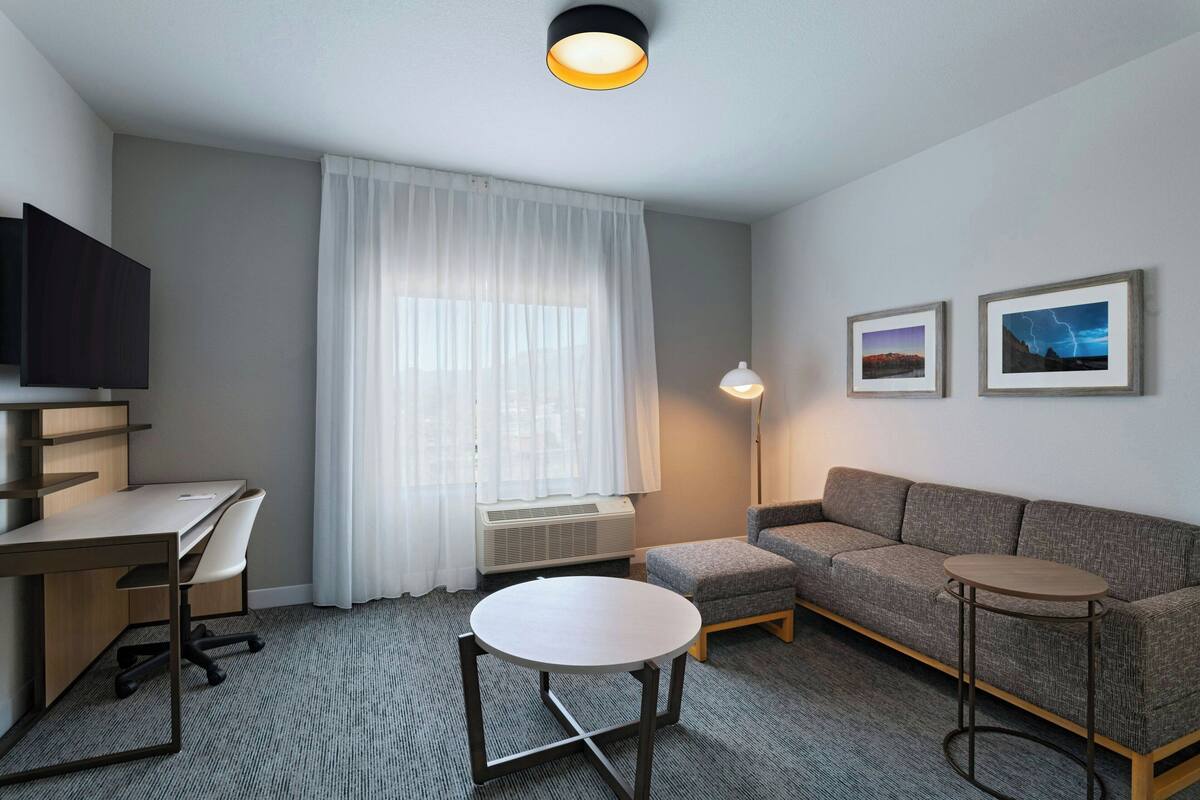 Une chambre d’hôtel équipée d’un bureau, de deux chaises, d’un canapé, d’une petite table ronde, d’un lampadaire et de deux tableaux encadrés accrochés au mur.