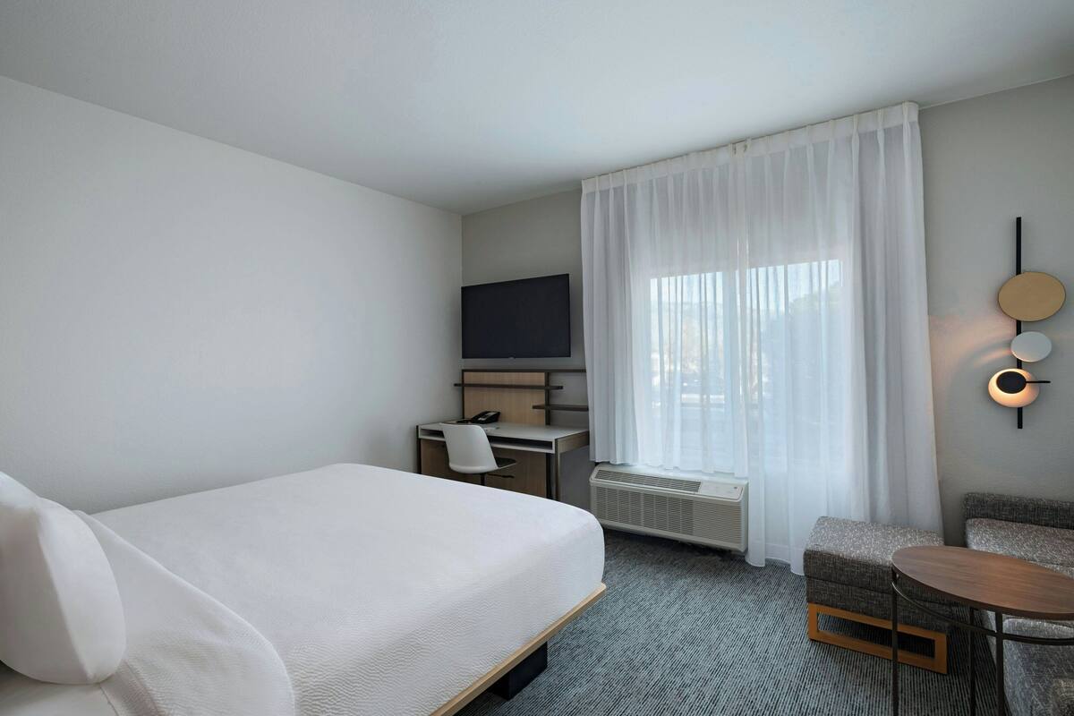 Une chambre d’hôtel comprenant un lit, un bureau avec une chaise, une télévision, une petite table et une fenêtre avec des rideaux.