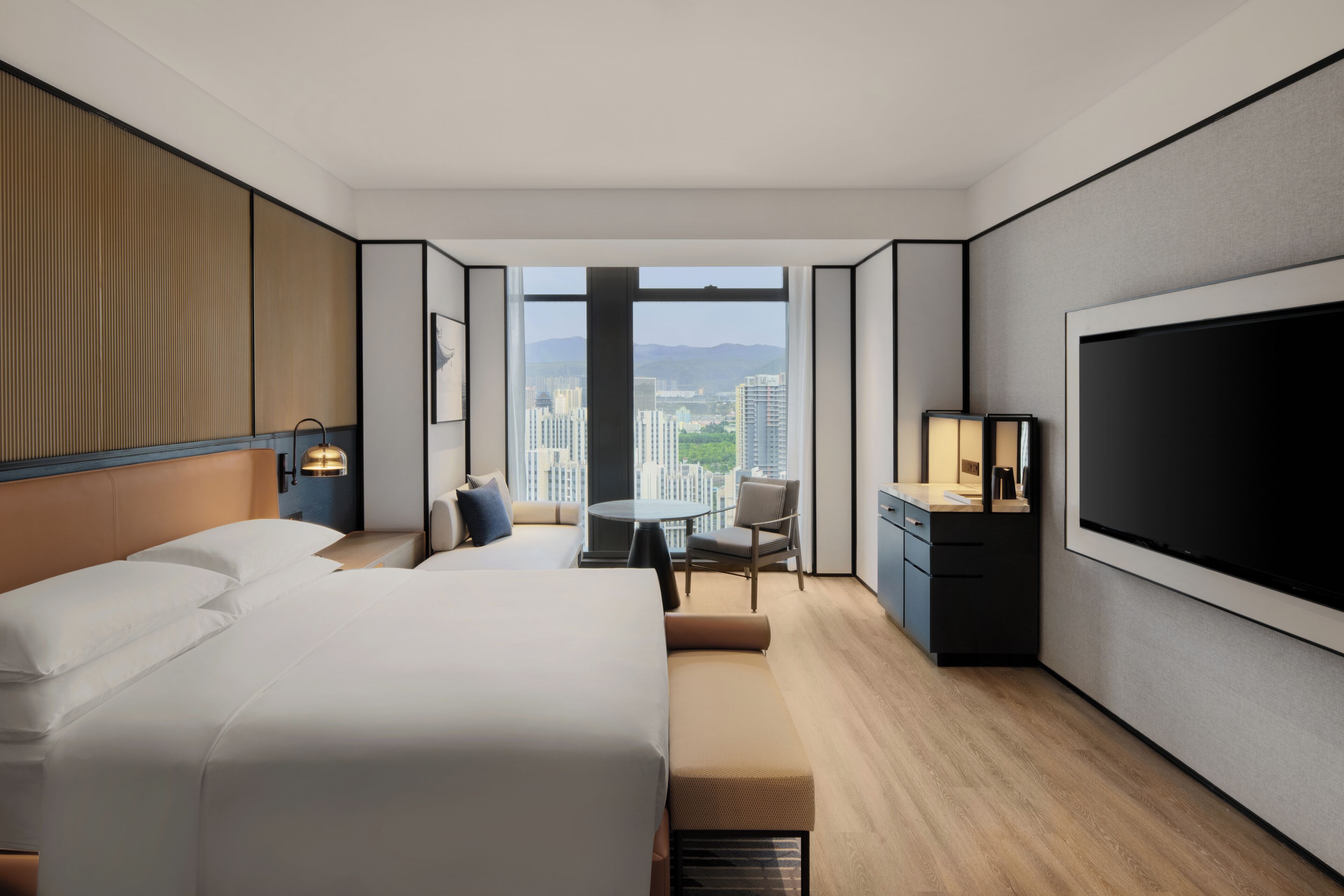 A modern hotel room with a large bed, a desk, a chair, a TV, and a view of the cityscape.