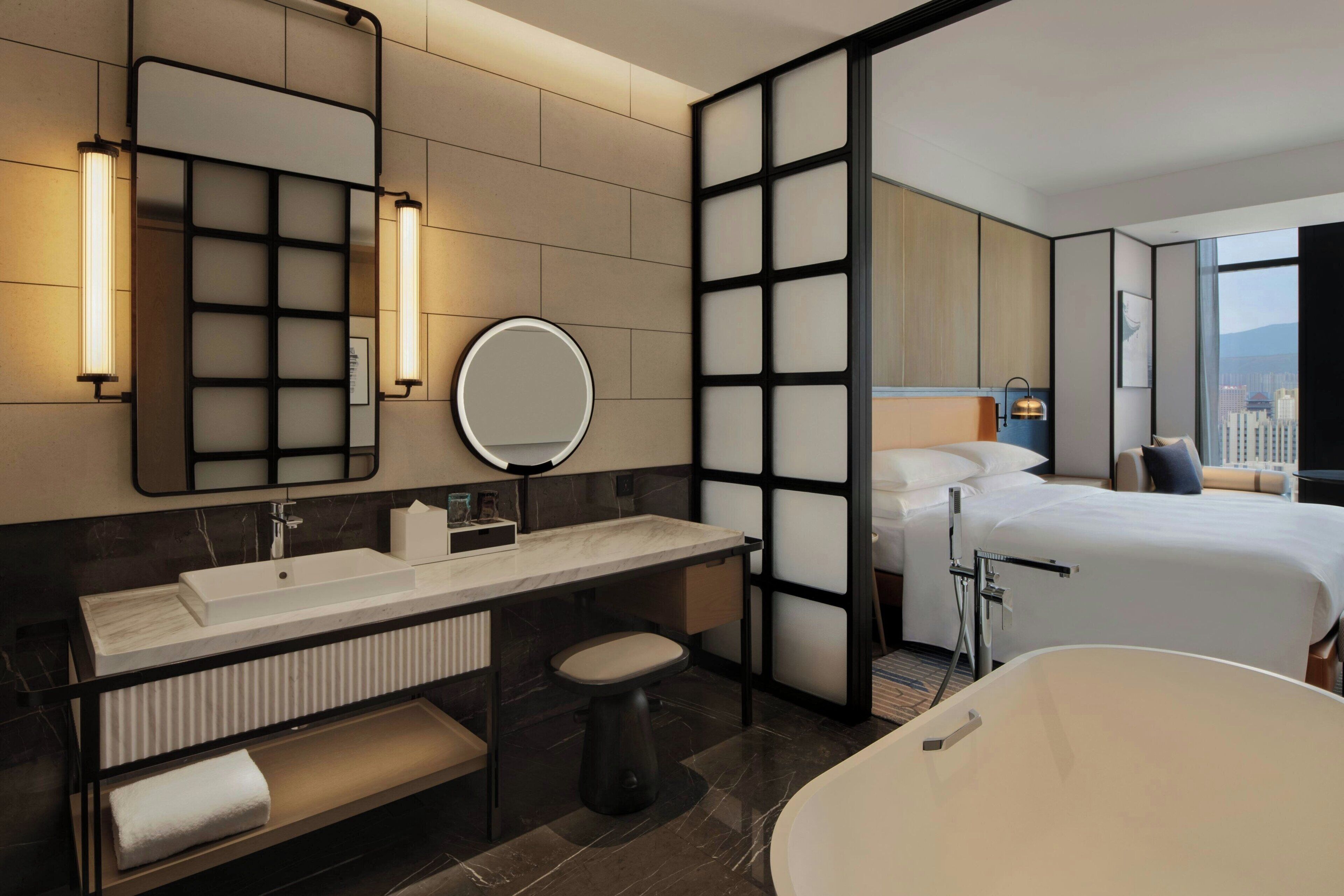 A modern hotel room with a large bed, a separate bathroom with a bathtub, a vanity with a mirror, and a city view through the window.