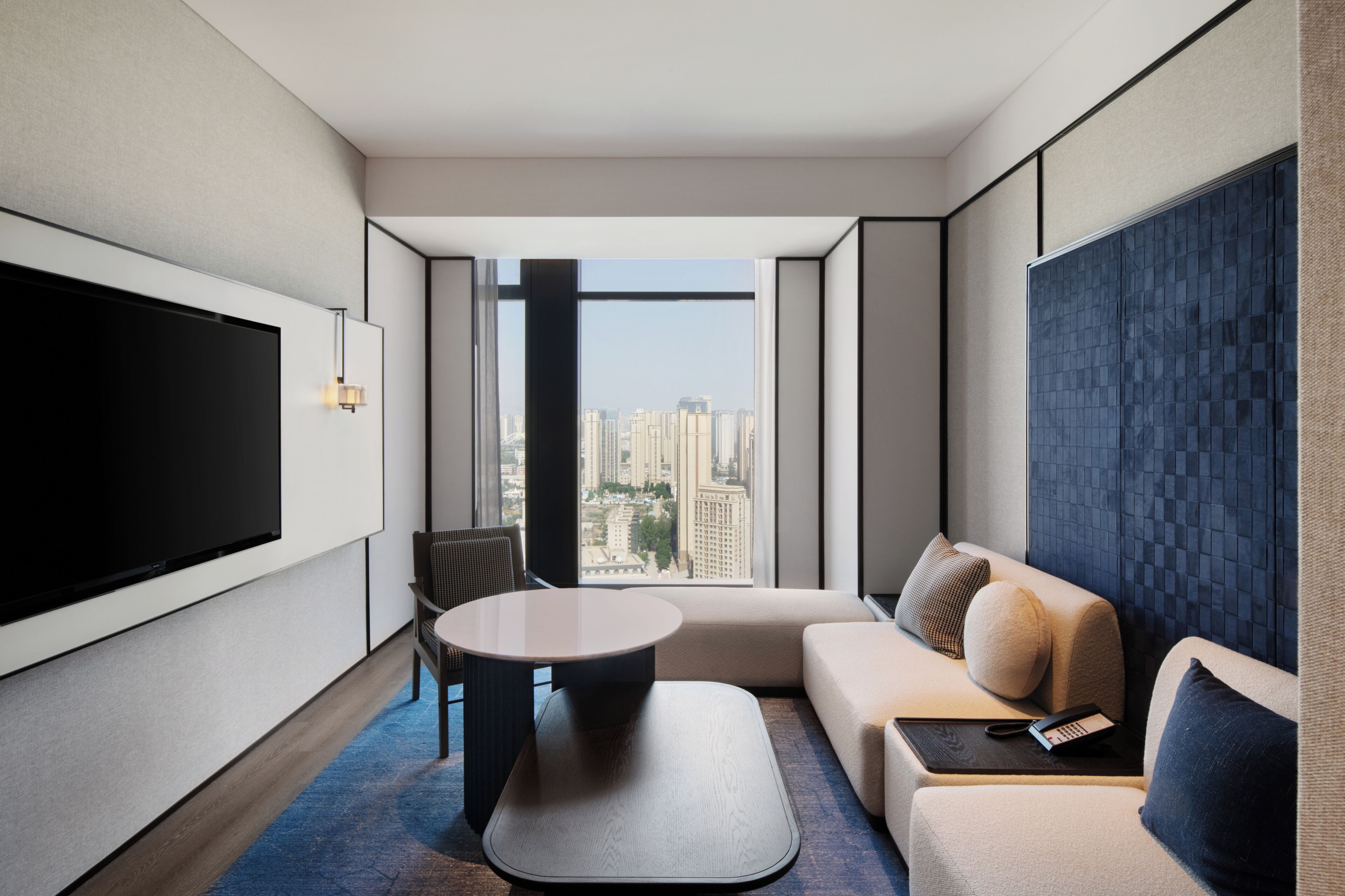 Una habitación de hotel moderna con un televisor de pantalla plana grande, un sofá, una mesita y vistas a la ciudad a través de amplios ventanales.