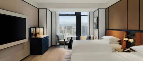 A modern hotel room with a large bed, a desk with a TV, a small table, and a view of the city through large windows.