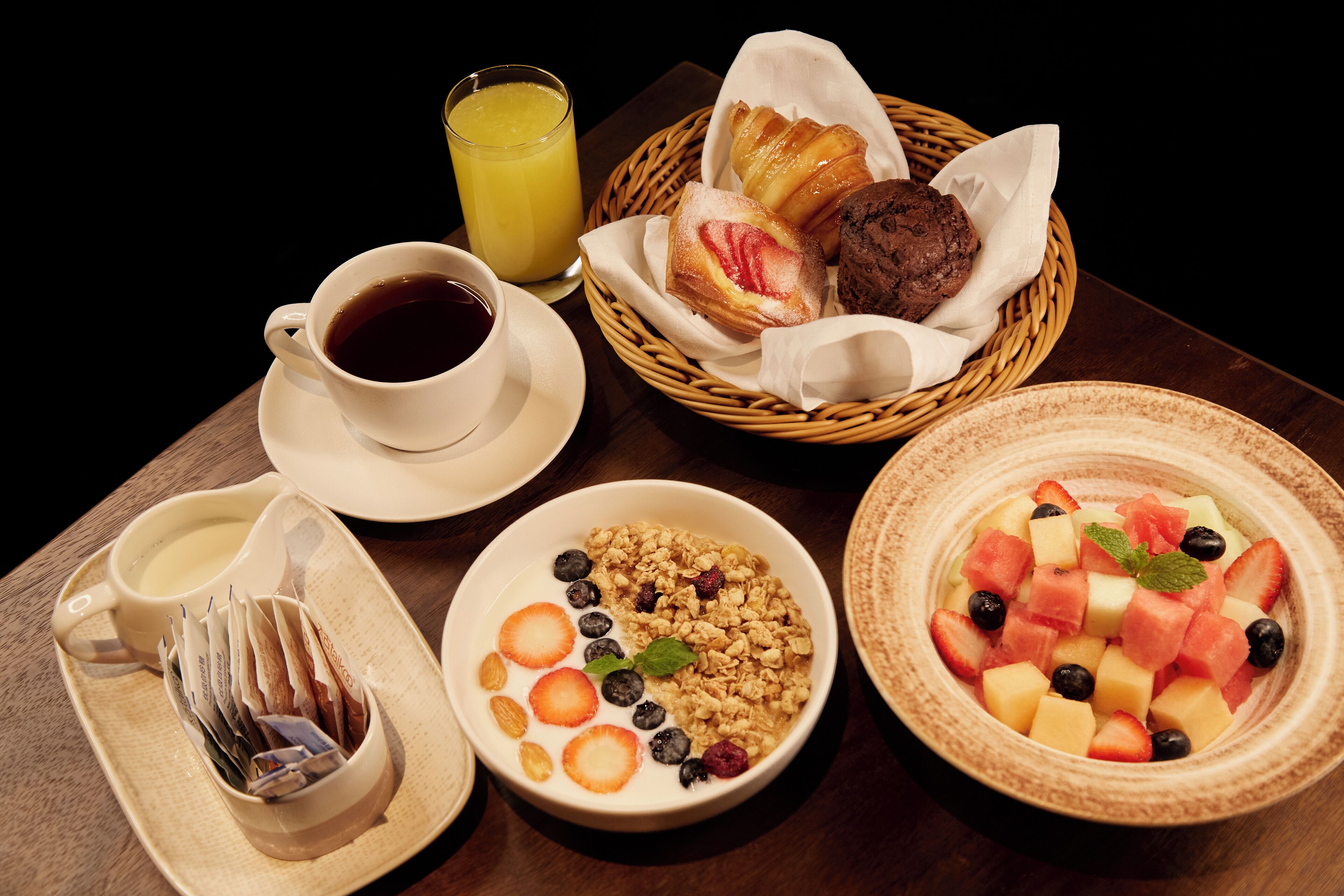 A breakfast setting with a cup of coffee, a glass of orange juice, a basket of pastries, a bowl of cereal with fruits, and a tray with a newspaper.
