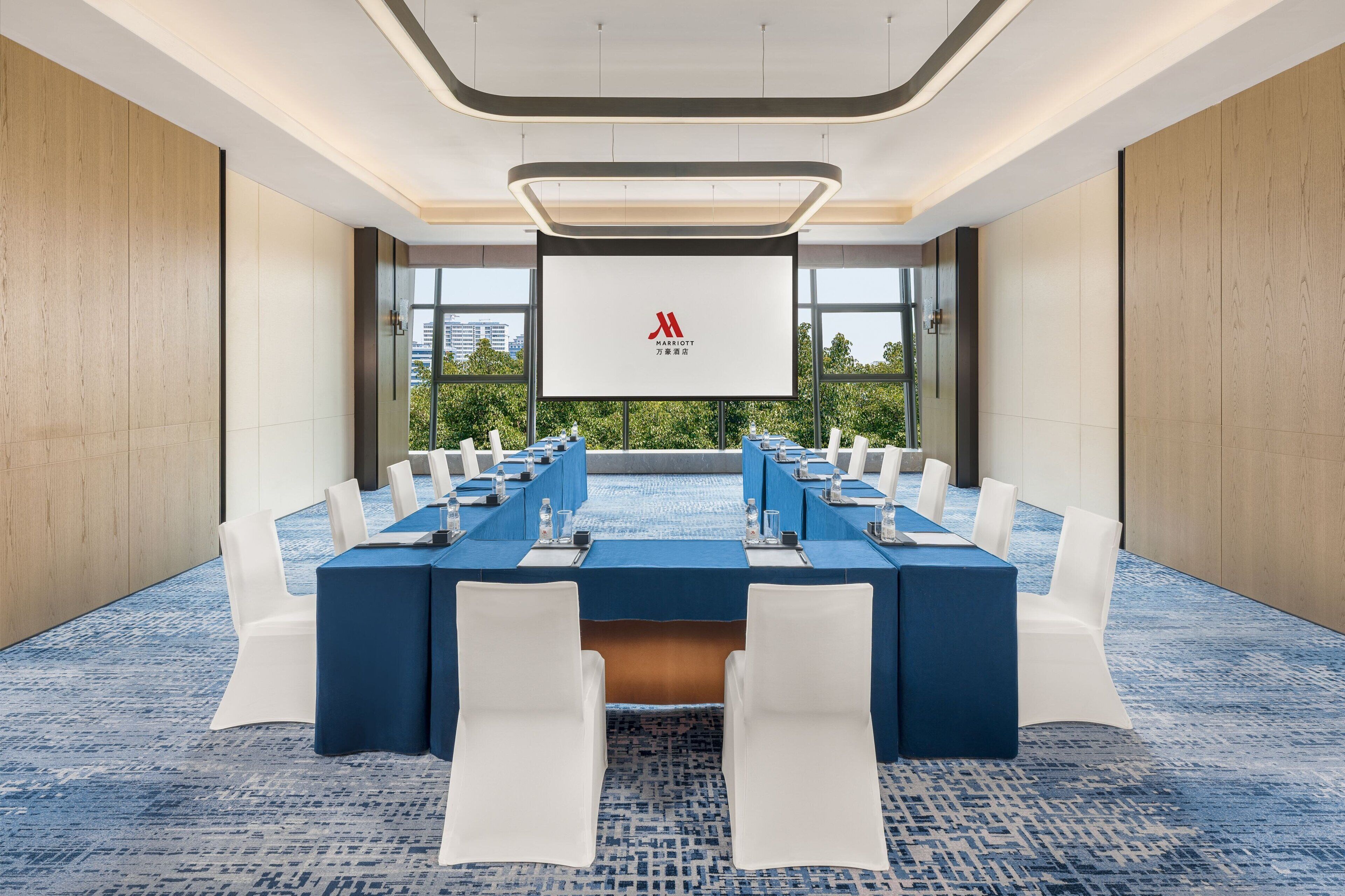 Een vergaderzaal met een lange tafel, witte stoelen en een groot scherm waarop het Marriott logo staat.