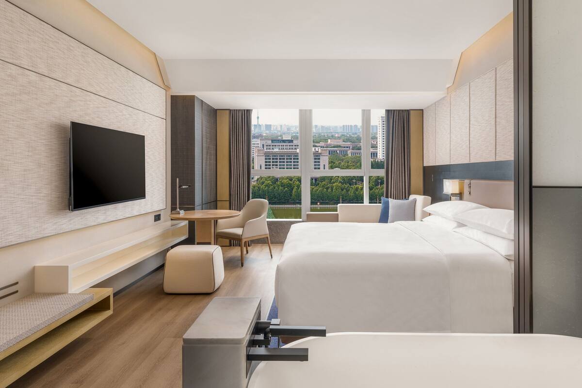Quarto de hotel moderno com uma cama grande, uma escrivaninha, uma cadeira e uma televisão na parede.