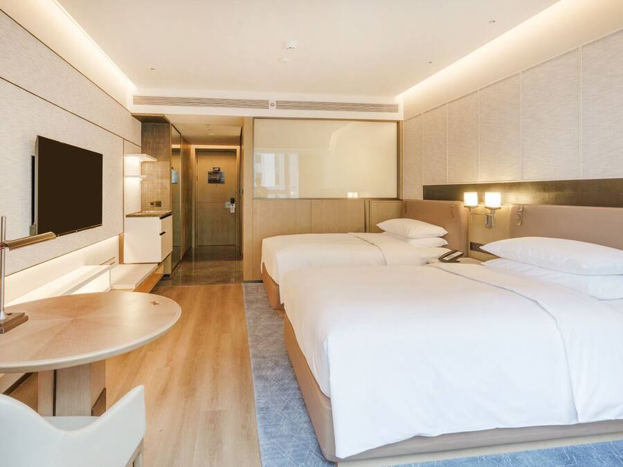A modern hotel room with a flat-screen TV, a desk, two beds, and a bathroom visible through a door.