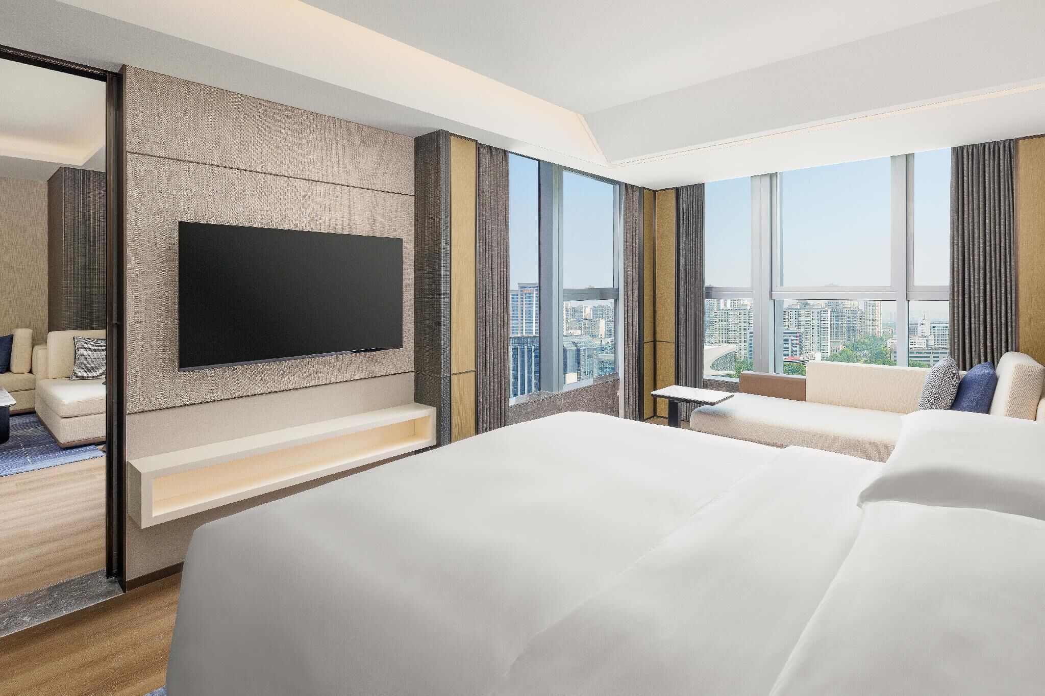 Una habitación de hotel moderna con una cama grande, un televisor de pantalla plana montado en la pared, un sofá y vistas a la ciudad a través de grandes ventanales.