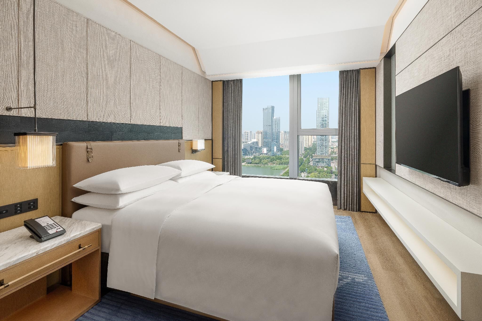Une chambre d’hôtel avec un grand lit, un téléviseur fixé au mur, une table de chevet avec un téléphone et une vue sur le paysage urbain.
