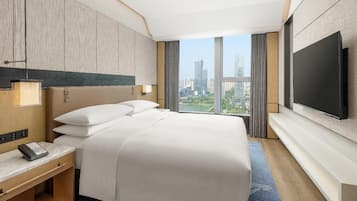 A hotel room with a large bed, a TV mounted on the wall, a bedside table with a phone, and a view of the cityscape.