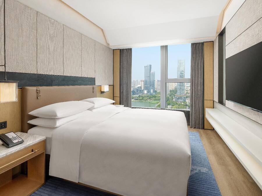 A hotel room with a large bed, a TV mounted on the wall, a bedside table with a phone, and a view of the cityscape.