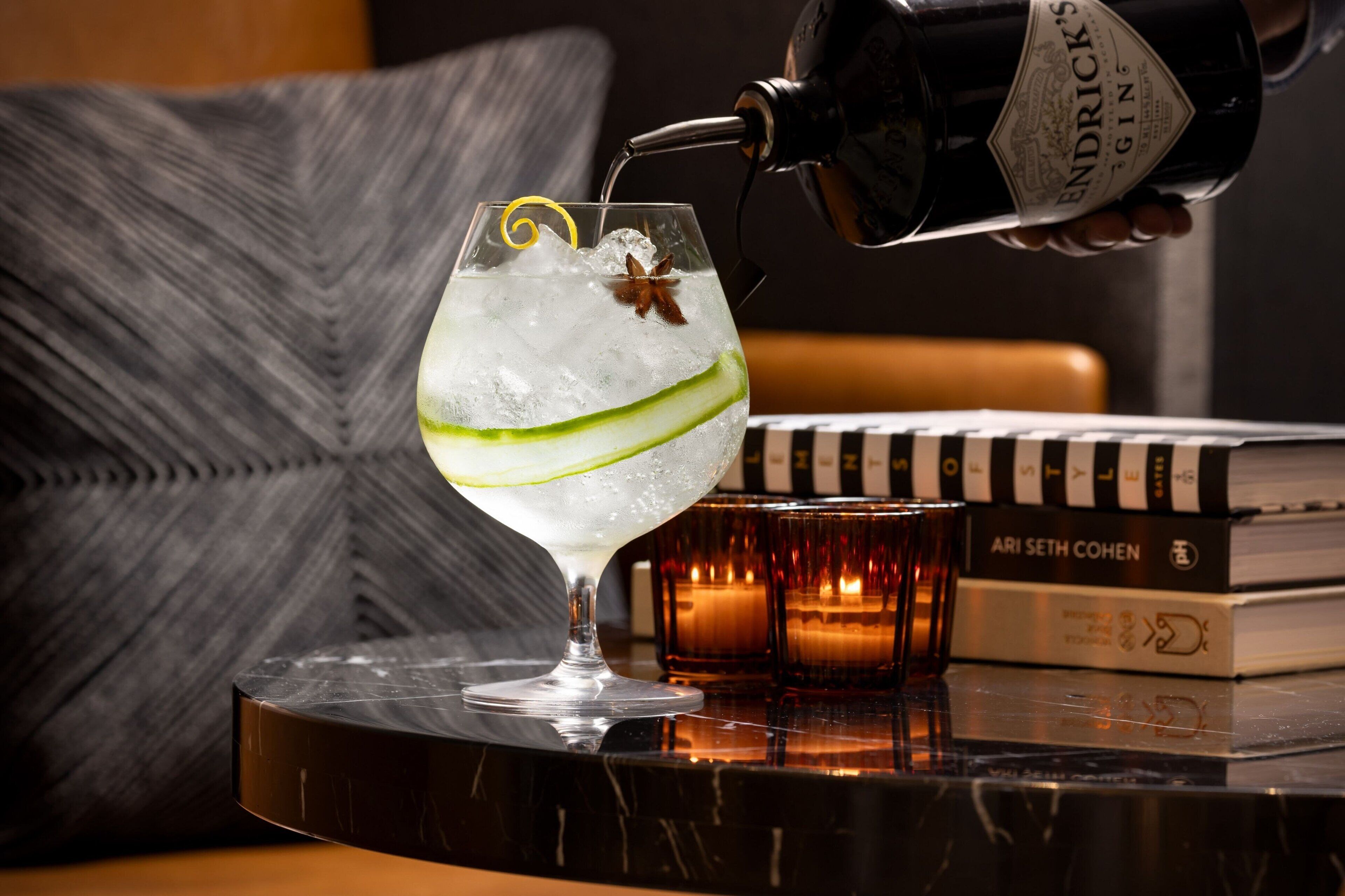 A cocktail being poured into a glass with a lime wedge and star anise, placed on a table with books and candles.
