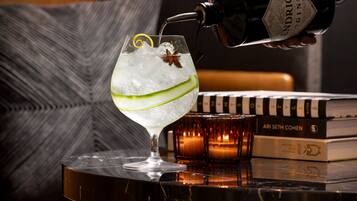 A cocktail being poured into a glass with a lime wedge and star anise, placed on a table with books and candles.