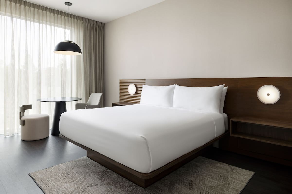 A modern hotel room with a large bed, a round table, a chair, and a small ottoman.