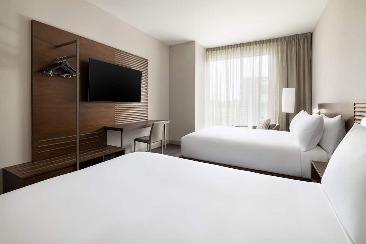 A hotel room with two beds, a TV mounted on a wooden panel, a desk with a chair, and a window with curtains.