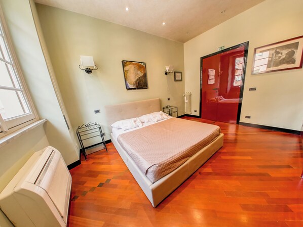 A hotel room with a bed, a radiator, a window, a door, and a wall-mounted light fixture.