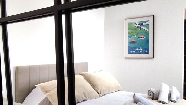 A modern hotel room with a bed, pillows, and rolled towels on the bed. There is a framed picture on the wall.
