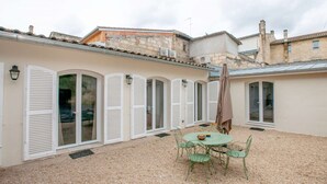A courtyard with a round table and chairs, white shutters, and a gravel ground.