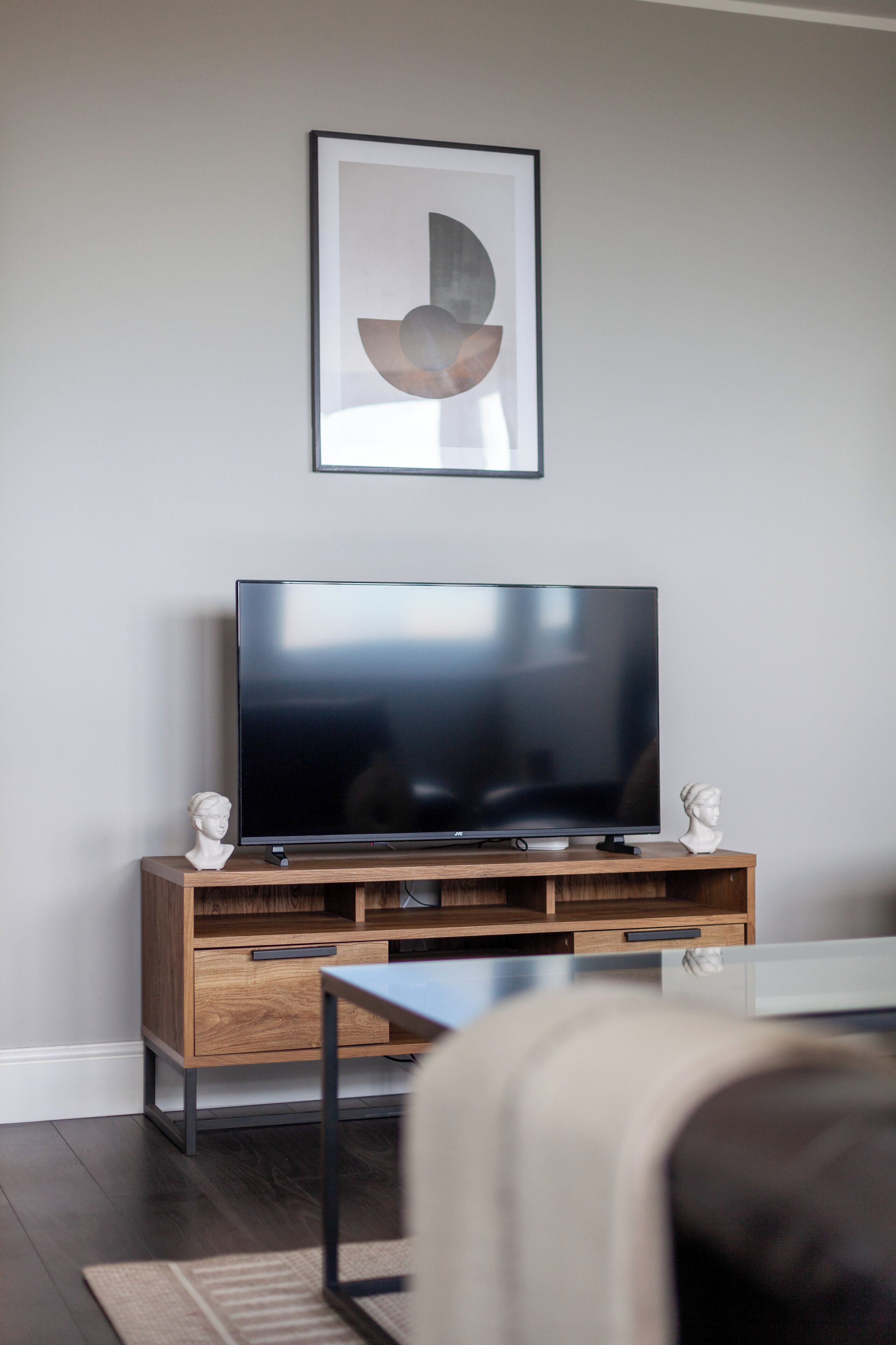 A modern living room with a flat-screen TV on a wooden stand, a framed abstract artwork above the TV, and a bust on either side.