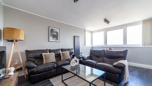 A modern living room with dark wood flooring, a glass coffee table, two black leather sofas, a wooden floor lamp, and a framed artwork on the wall. - The Old Street Luxury Apartment (London)