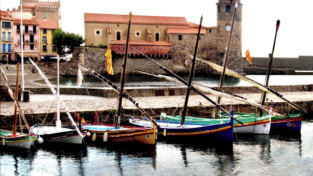 A harbor with several colorful boats docked, a stone building with a tower, and a flagpole with a flag. - Le Nid de Gilles (Collioure)