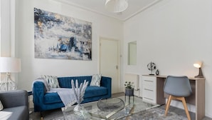 Uma sala de estar moderna com um sofá azul, uma mesa de centro de vidro, uma escrivaninha com uma cadeira e um quadro abstrato grande na parede.