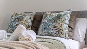 Cama arrumada com um cobertor verde e duas almofadas com padrão de folhagem tropical.