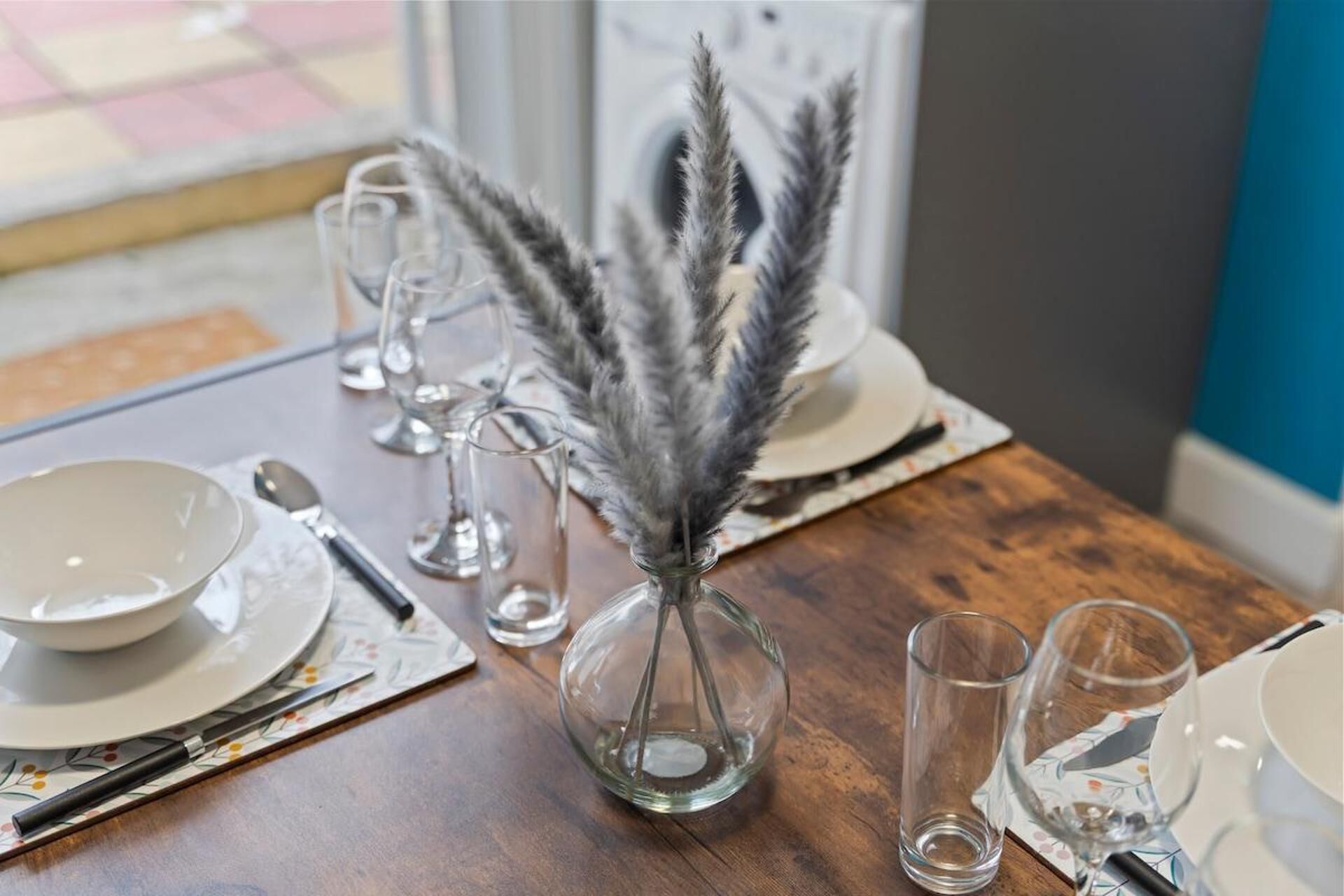A dining table with a clear glass vase containing a feather arrangement, a set of cutlery, and several empty glasses.