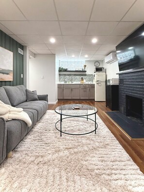 A living room with a grey sofa, a glass coffee table, a fireplace, and a kitchen in the background.