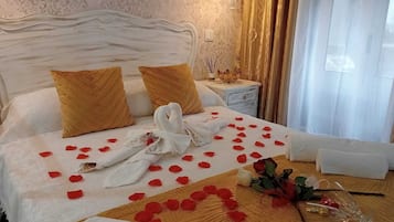 A neatly made bed with rose petals and a heart-shaped arrangement, a white headboard, and a bedside table with a lamp and a bowl.