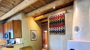 A cozy living room with a fireplace, wooden ceiling beams, a sofa, a coffee table, and a kitchen area with a microwave. - Sweet Seco Casita is a classic Taos Adobe Home (Arroyo Seco)