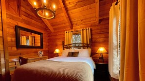 Un dormitorio acogedor tipo cabaña de madera, con cama, dos lámparas de noche, un espejo y una ventana con cortinas.