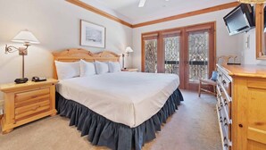 A bedroom with a large bed, wooden headboard, two bedside tables with lamps, a ceiling fan, a TV mounted on the wall, and a view of the outside through a glass door.