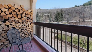 A balcony with a stack of firewood, a metal chair, and a view of a residential area with trees and a building.