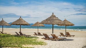 A beach with thatched umbrellas and wooden lounge chairs. - Abogo Da Nang OceanFront Luxury Villa (Da Nang)