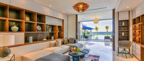 A modern living room with a sofa, ottoman, and coffee table, wooden shelves with decorative items, and a view of the beach through large windows.