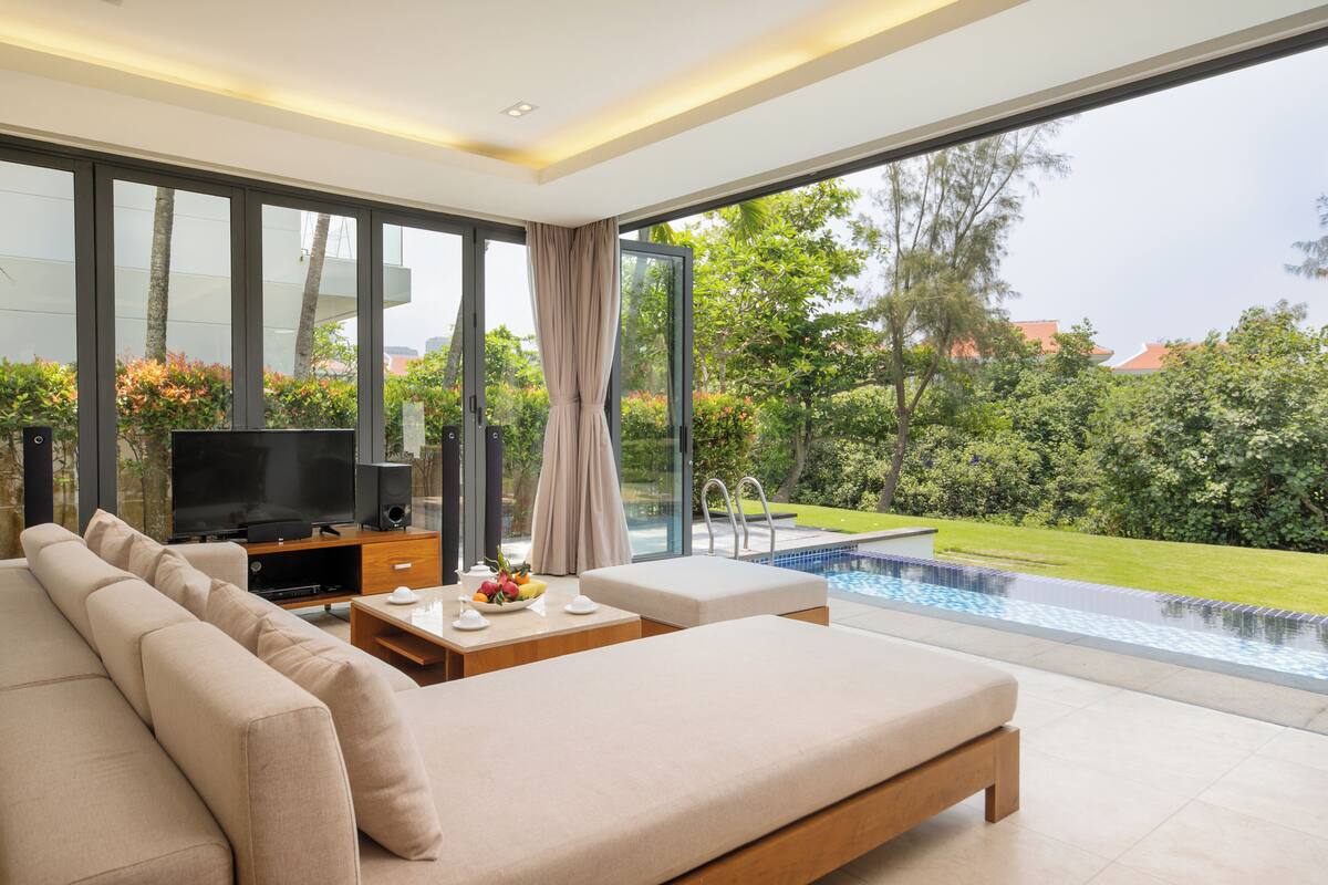 Una sala de estar moderna con un televisor grande, un sofá y una mesa de comedor con fruta. Se ve una piscina a través de los amplios ventanales.