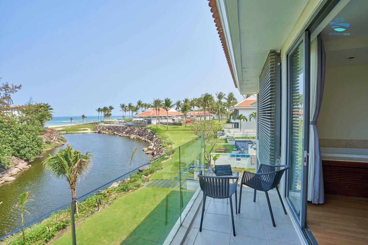 Un balcón con vista a un río y palmeras, una computadora portátil sobre una mesa y una silla.