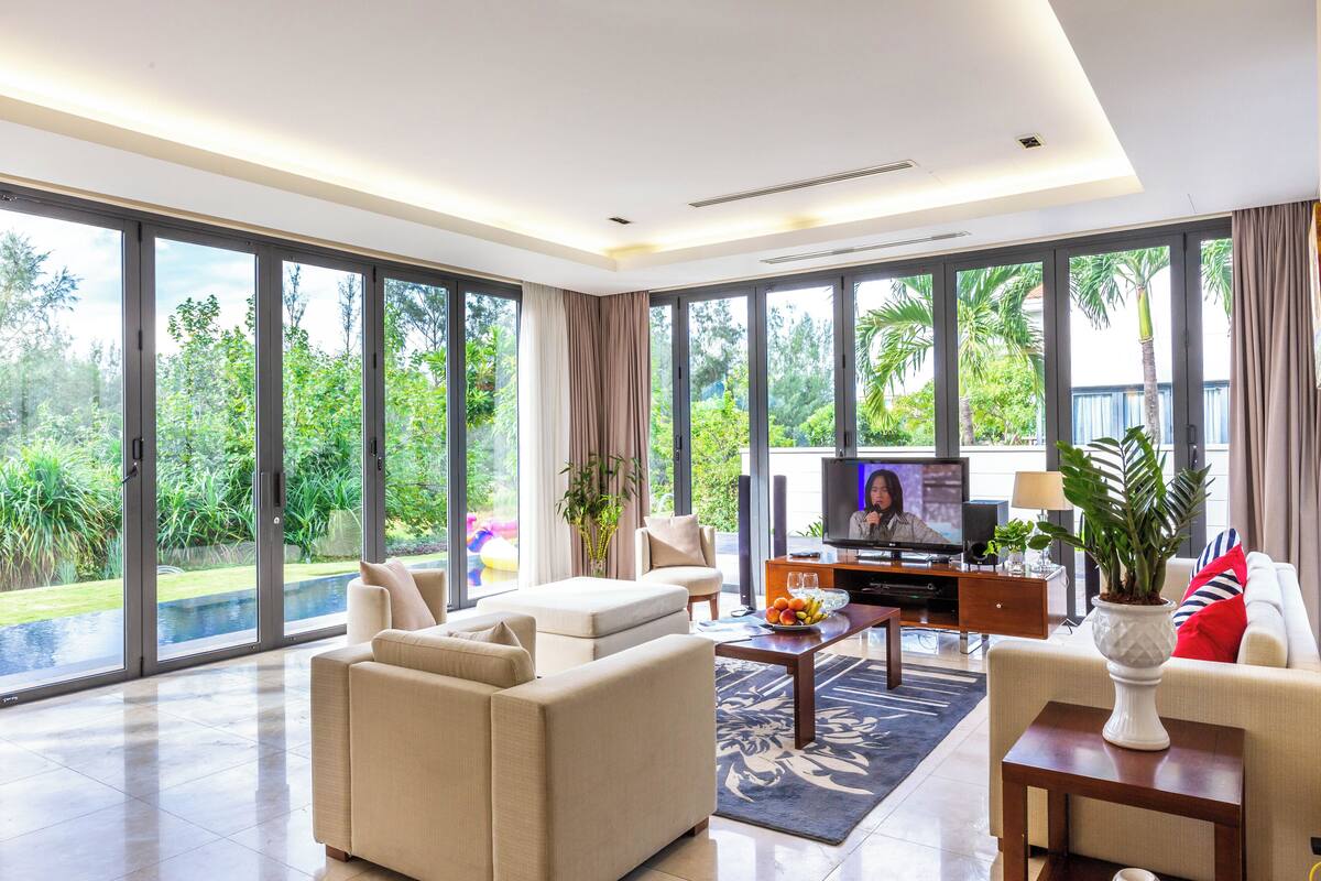 Amplia sala de estar con televisor grande, asientos cómodos y vista a un jardín exuberante a través de amplias ventanas.