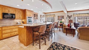 A spacious kitchen with wooden cabinets, a central island with bar stools, a dining area with a table and chairs, and a living room with a sofa and a lamp. - The Charter at Beaver Creek F3505 (Beaver Creek)