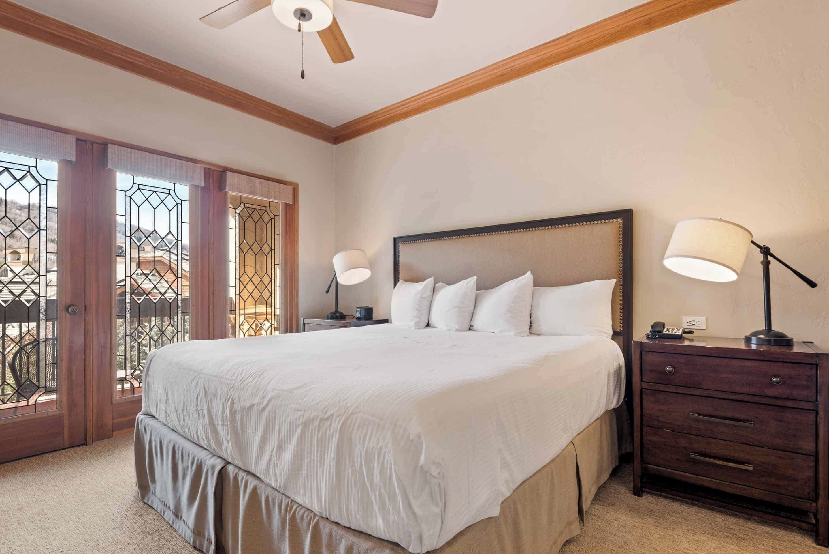 A bedroom with a large bed, wooden headboard, two bedside lamps, a nightstand, and a view through a glass door.
