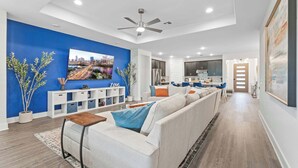 A modern living room with a large sofa, a coffee table, and a TV. There is a kitchen in the background with cabinets and a refrigerator. - Close to CotA & Downtown | Spacious & Luxe | 4BR (Austin)