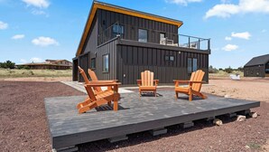 A modern two-story house with a balcony, wooden deck with chairs, and a clear sky. - *NEW* Luxe Desert Retreat | Near GrandCanyon S Rim (Williams)