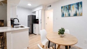 A modern kitchen with white cabinetry, a stainless steel sink, and a black faucet. There is a dining area with a round wooden table and white chairs. A painting is on the wall, and a plant is on the table.