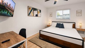 A bedroom with a bed, a desk, a chair, a ceiling fan, artwork on the walls, and a window.