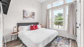A bedroom with a large bed, a side table with a lamp, a ceiling fan, a window with curtains, and a painting on the wall. - Close to CotA & Downtown | Cozy & Luxe | 4BR (Austin)