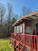 Una cabaña pequeña con fachada roja, una terraza de madera y una ventana con vista a un área verde.