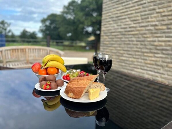Una mesa con diversos alimentos, como pan, queso, frutas y huevos, junto con dos copas de vino tinto.
