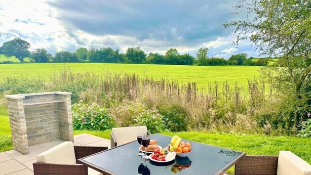 Eine Terrasse im Freien mit einem Tisch, der für eine Mahlzeit gedeckt ist, umgeben von Korbstühlen, mit Blick auf ein grünes Feld.