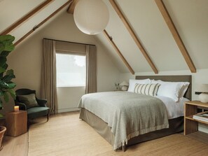 A bedroom with a sloped ceiling, a large window with curtains, a bed with multiple pillows, a bedside table with a lamp, and a potted plant. - The Great Bustard (Salisbury)